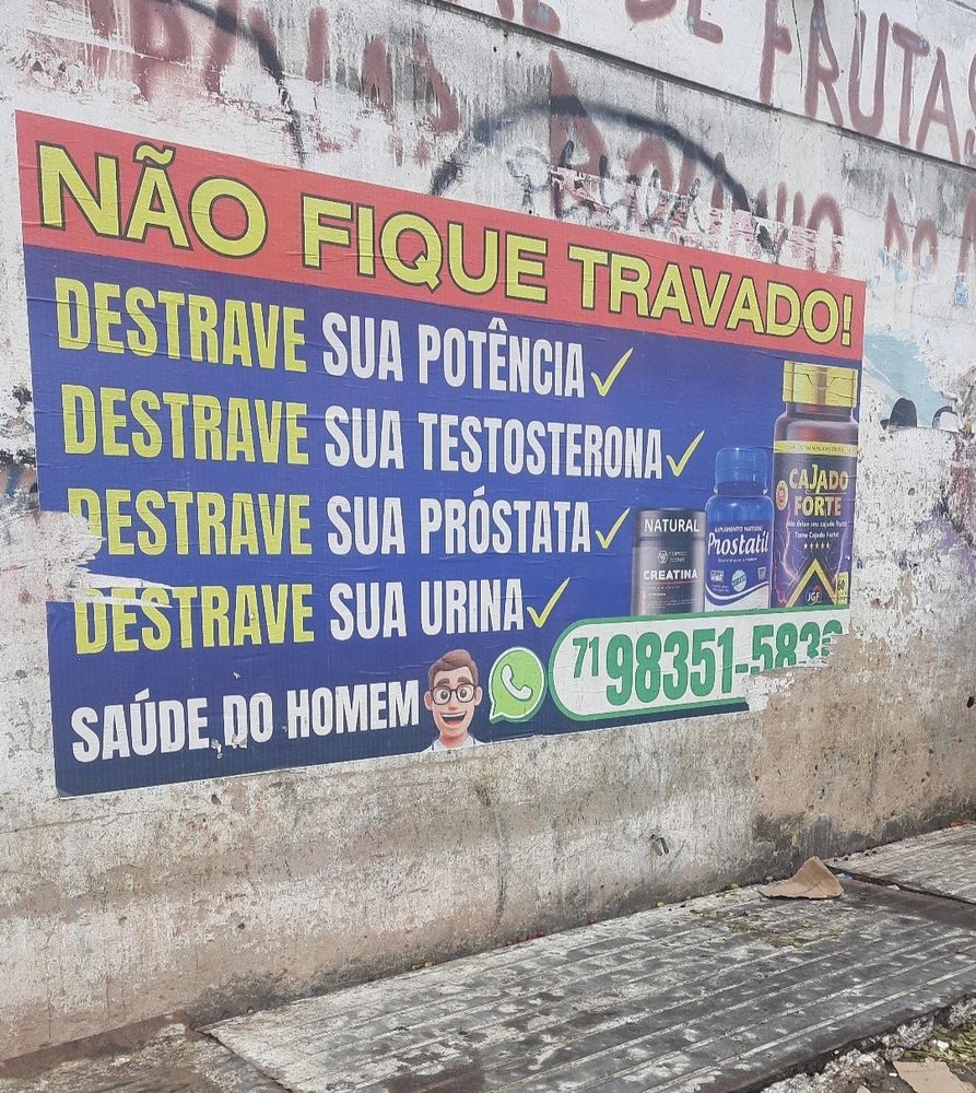 Cartaz de anúncio colado em um muro pixado. Uma faixa na parte de cima, com o fundo vermelho e as letras amarelas, dizendo: não fique travado! 
Em baixo, em um fundo azul, com letras amarelas e brancas: Destrave sua potência ✔️ Destrave sua testosterona ✔️ Destrave sua próstata ✔️ Destrave sua urina ✔️ Saúde do homem. Ao lado das frases, 3 embalagens de suplementos: Creatina, Prostatil e Cajado Forte.
Um desenho de um homem em 3D e o símbolo do Whatsapp com um número de telefone ao lado. 