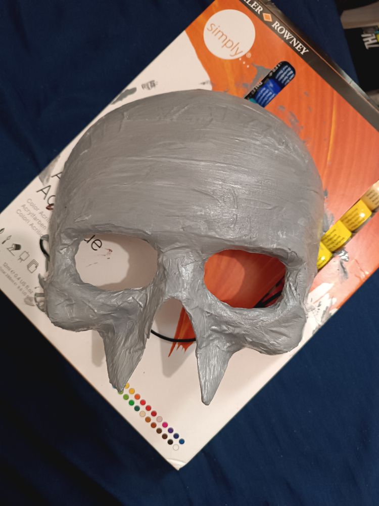 Uma máscara de caveira pela metade, até o nariz, pintada de cinza prateado. Ela está repousando sobre uma caixa de tinta acrílica. 