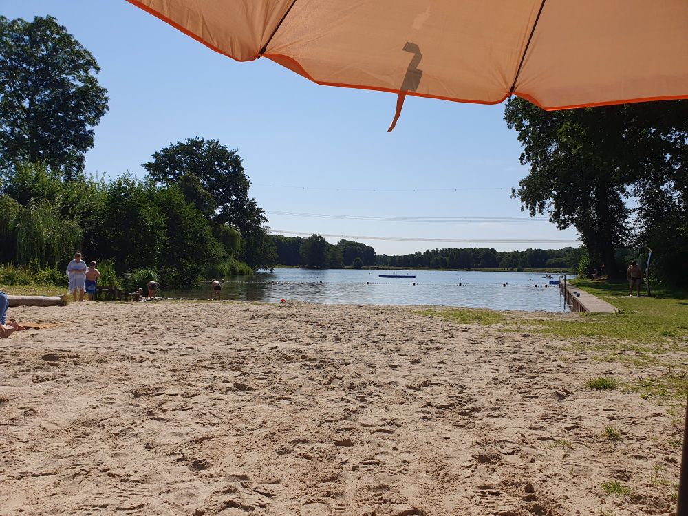 Blick auf einen kleinen See aus liegender Position. Am oberen Bildrand der Rand eines Sonnenschirms. Im Vordergrund Strandsand. Der See ist umrandet von vielen Büschen und Bäumen. Der Himmel ist wolkenlos blau.