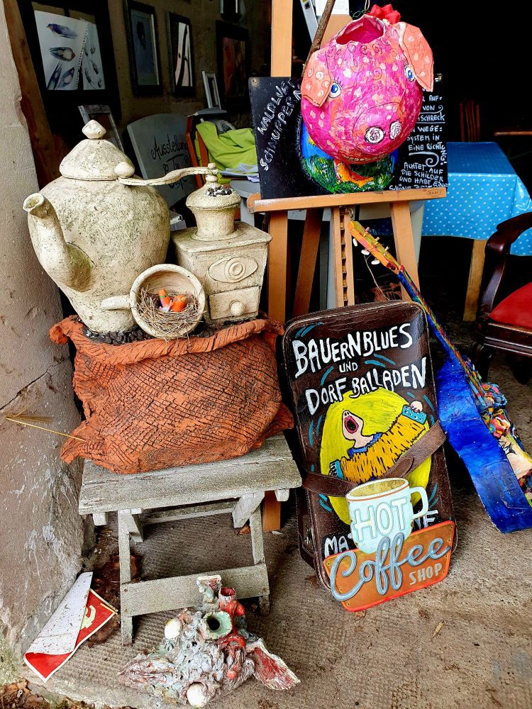 Sammelsurium in der Scheune einer Künstlerin: Getöpfertes aus weißem Ton, z.B. Eibe Kaffeekanne und Kaffeemühle; ein selbstgemachtes buntes Schild für eine Musikveranstaltung, eine blaue Gitarre, ein pinkes Schwein aus Pappmachee usw.