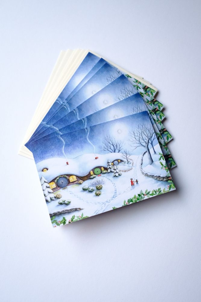 Image shows a stack of four cards with my illustration “A Very Snowy Hobbiton” on it. The painting includes a series of three hobbit homes under a single hill. They are blanketed with snow and cheerful light spills from their windows. A full moon shines over a blue evening sky. In the foreground, a tiny hobbit mom and child are walking to one of the homes. 