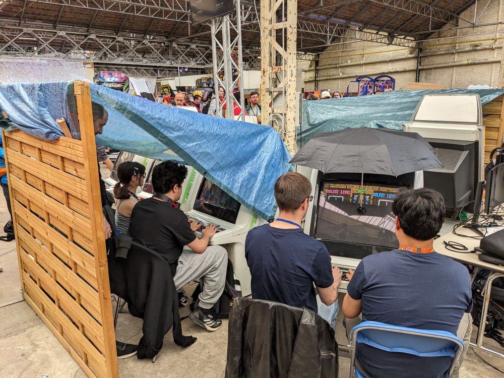 Les bornes d'arcade sur lesquels se jouent le tournoi Puyo Puyo 2, avec un parapluie et une bâche accroché à un panel en bois pour éviter les reflets de lumière sur les écrans 