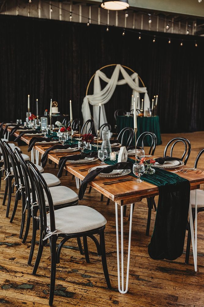 Farm table with green velvet table runner, scattered flowers in bud vases, white plates and black napkins. Black bentwood chairs and a black velour backdrop 