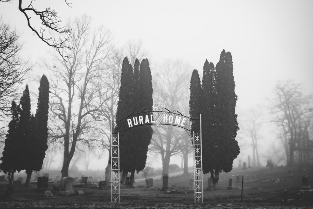Entrance sign to the Rural Home Cemetery in Vernon, Wisconsin. There are gravestones and trees obscured by a cool November fog. 