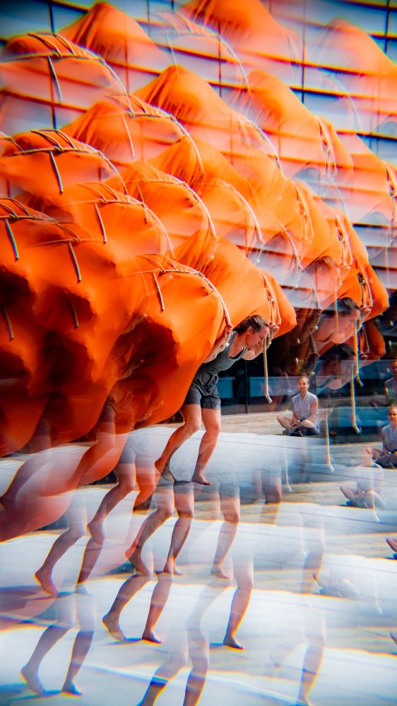 A picture of a woman in grey clothing carrying a large orange bundle. The photograph has an effect on it which repeats the image and makes it look like you're looking at it through a kaleidoscope.