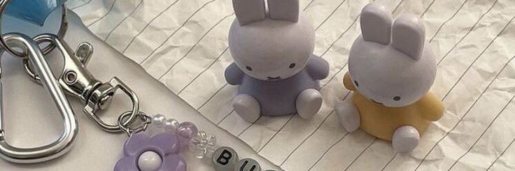 Image of two miffy bunnies on a crumpled piece of paper. One miffy bunny is in a lavender dress while the other is in a soft yellow one. Next the the bunnies on the paper is a silver keychain attached to a carabiner - it has a purple flower charm and a beaded word charm that only has the fist couple beads shown: two white pearl beads, a lilac bead, a see through flower bead, a white and black “B” bead and a white and black “U” bead.