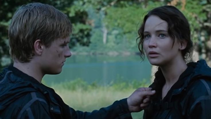 Katniss Everdeen and Peeta Mellark from The Hunger Games (2012). They’re in a forest with a lake in the background. They’ll both in solid black outdoor wear. Peeta (played by Josh Hutcherson) is blonde. He is entirely focused on Katniss’ face. He has a few scratches on his face and is reaching out gently touching Katniss’ braid in front of him. Katniss (played by Jeniffer Lawrence) is looking out past the camera, fear on her face. She has long brown hair that has been braided over her right shoulder.