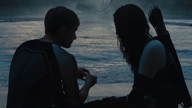 Katniss and Peeta from Catching Fire (2013). They’re on a man made beach in the hunger games area. You can see gentle waves behind them as well as a large cornucopia-shaped structure made of stone in the far distance. The lighting is blue. Peeta is turned to face Katniss. He has an oyster with a silver pearl in his hands. Katniss similarly faces Peeta though her haze is downwards towards what’s in his hands. Her long hair is out and reaches down her chest. They’re both silhouetted, you cannot make out facial features but you can see the outfits they wear are mostly dark water suits with long sleeves that are white from the shoulder to the mid arm. Katniss has a sheath full of arrows on her back.