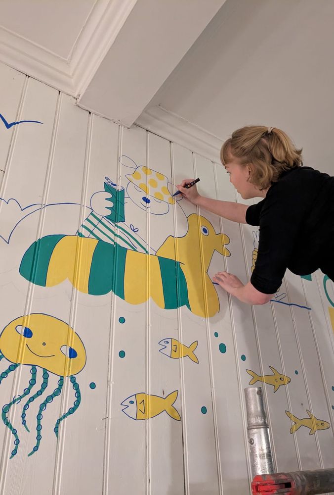 me drawing a yellow and green mural on a white wall with fish and a dog relaxing on a floating rubber duck
