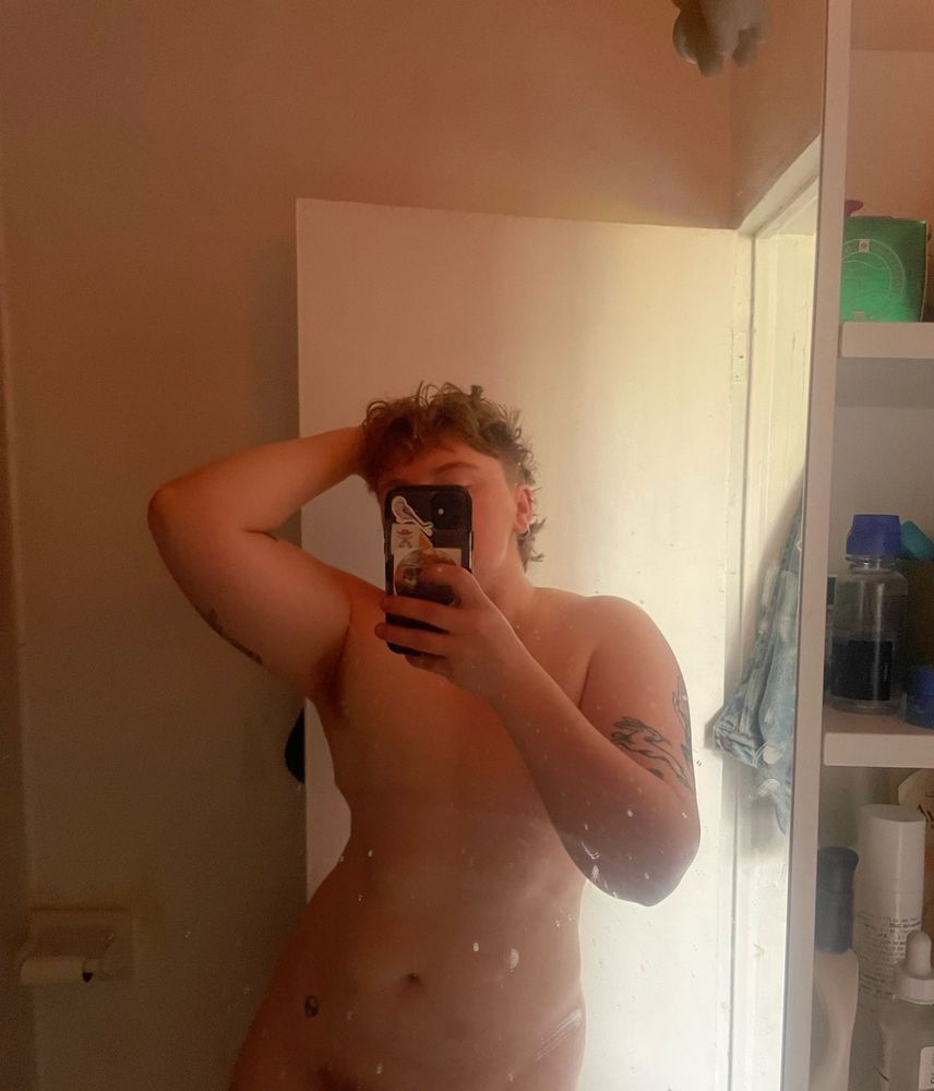 white trans masc person taking a mirror selfie. they are naked and showing off their tattoos and chest 