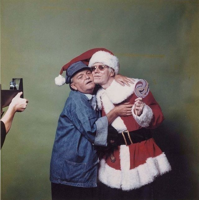 Andy Warhol dressed as Santa, holding a lollipop, hugging (or being hugged by) Truman Capote, in a studio. On the left is someone holding a camera, taking pictures of them