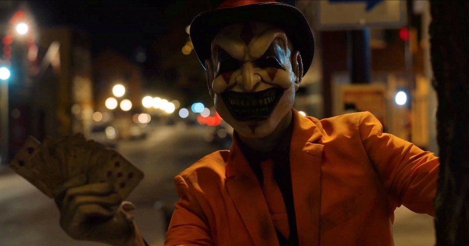 A creepy clown guy in an orange suit holds a deck of cards, on a street at night