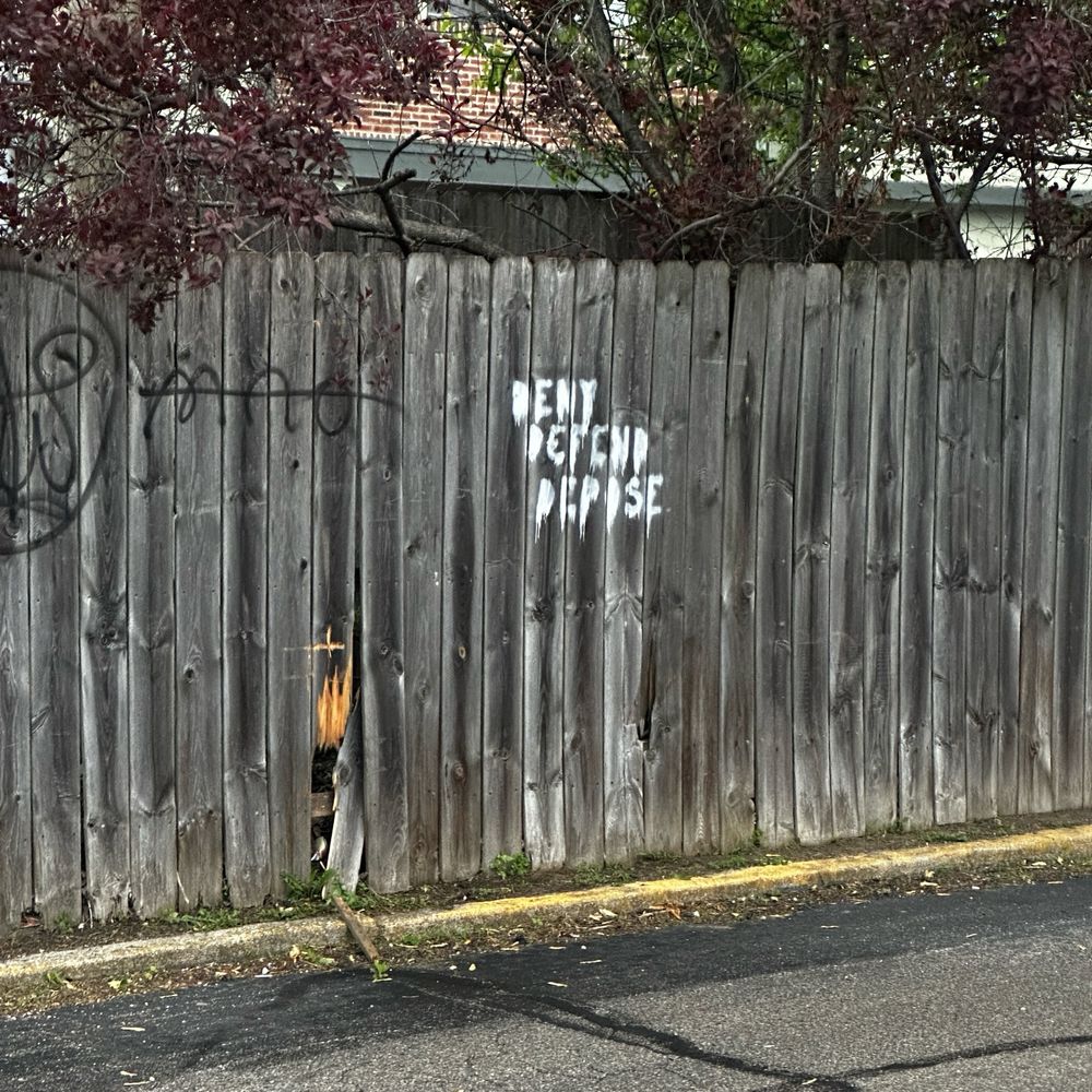 Picture of a wooden fence with “deny, defend, depose” spray-painted in white. Older graffiti is to the left of an abstract face with “MAO” in black paint