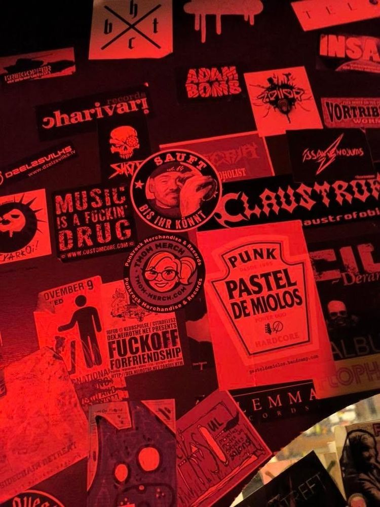 A wall covered with punk and music stickers. Right in the middle the sticker from tmom-merch.com 