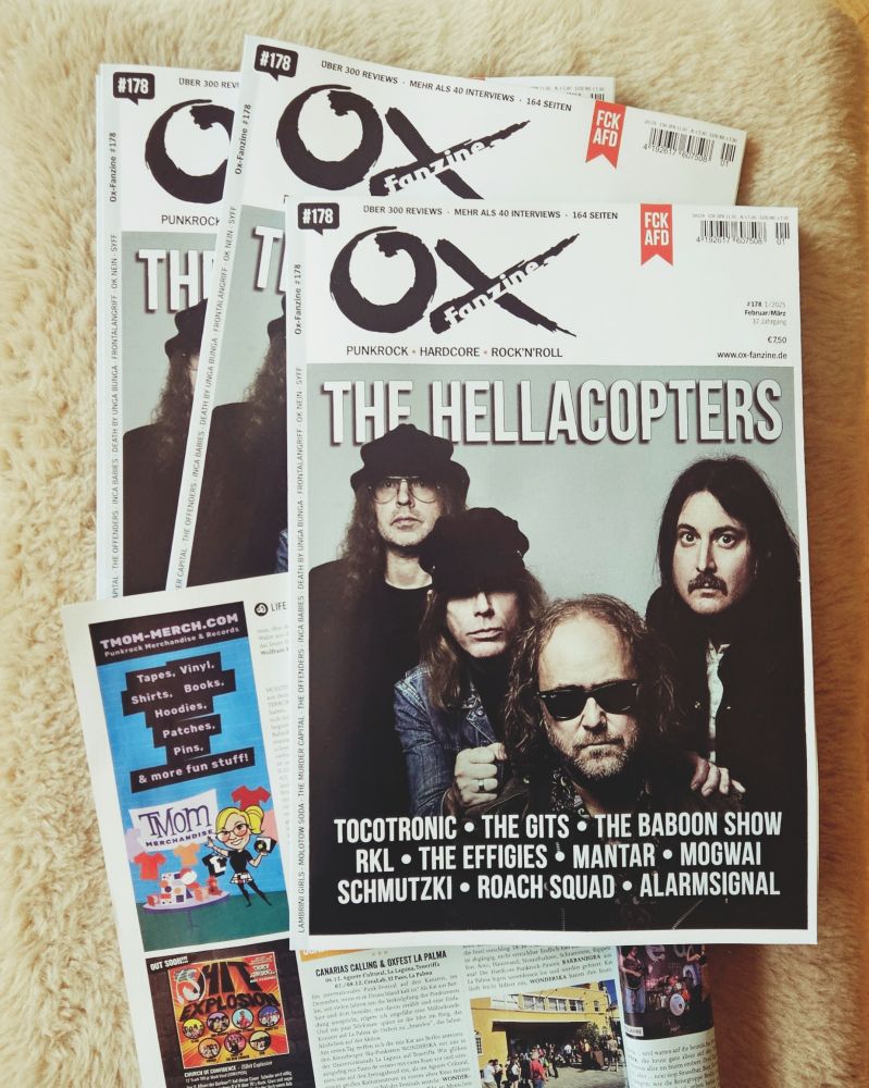 Mehrere Exemplare des neuen Ox Fanzine liegen übereinander. Eine Seite ist aufgeklappt und man sieht die Anzeige von TMom-Merch.com mit dem Text "TMom-Merch.com, Punkrock Merchandise & Records. Tapes, Vinyl, Shirts, Books, Hoodies, Patches, Pins & more fun stuff!" 