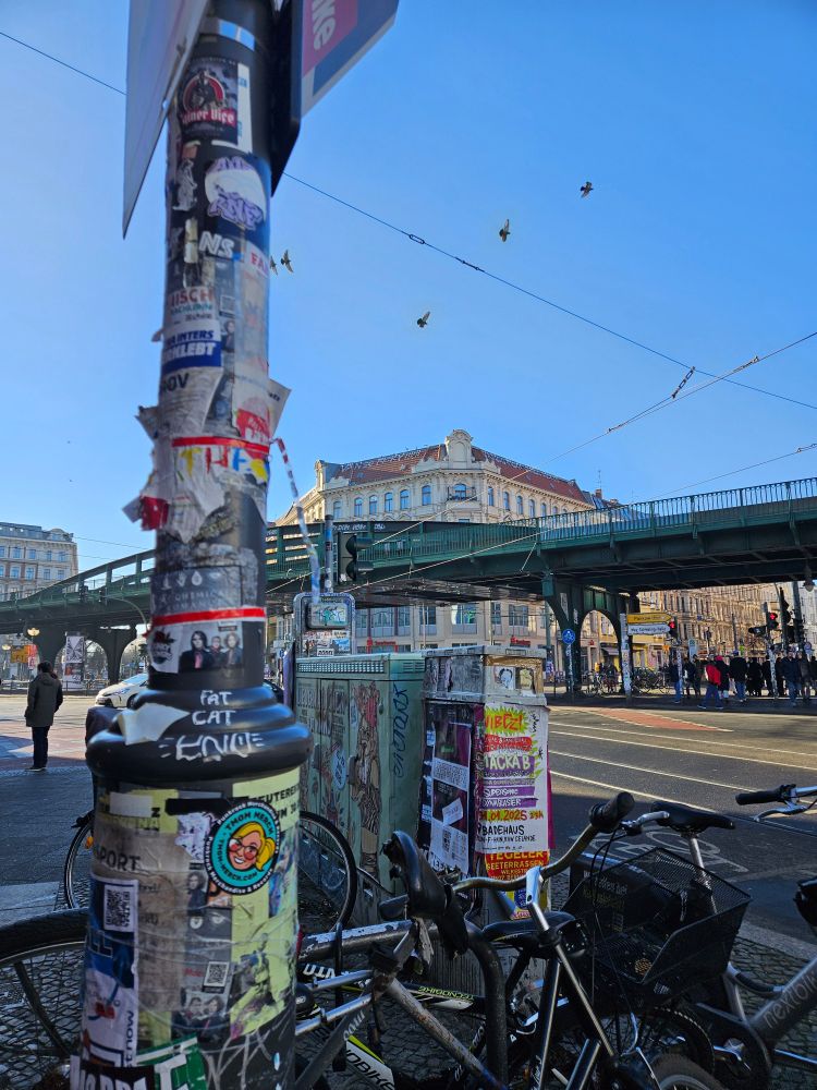 A busy street intersection in Berlin, Germany on a sunny day. In the front is a pole covered with poster, flyer, sticker and graffiti. One sticker is from the company TMom Merchandise. 