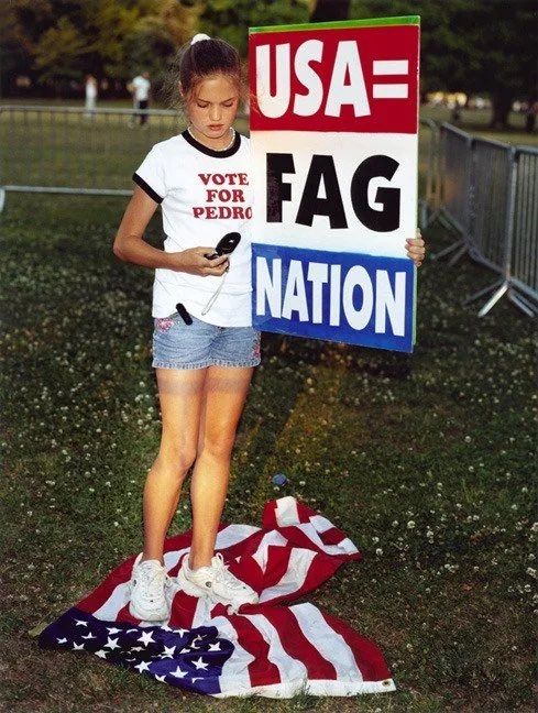 A teen girl wearing a Vote for Pedro t shirt looks at her flip phone while holding a placard that reads "USA=FAG NATION" and standing on a trampled American flag 