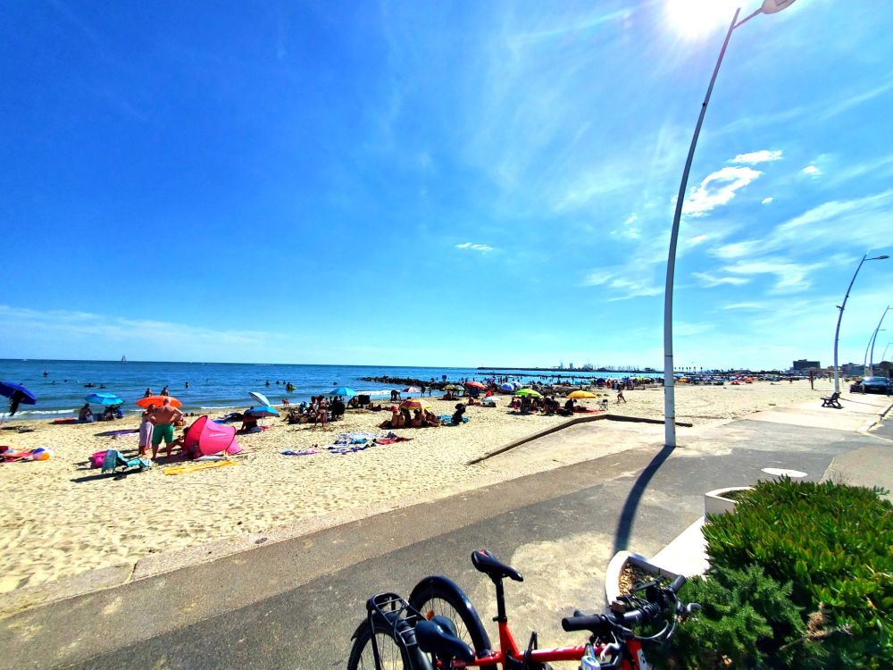 Beach with people and umbrellas. Bike path in the front with some bikes locked up. 