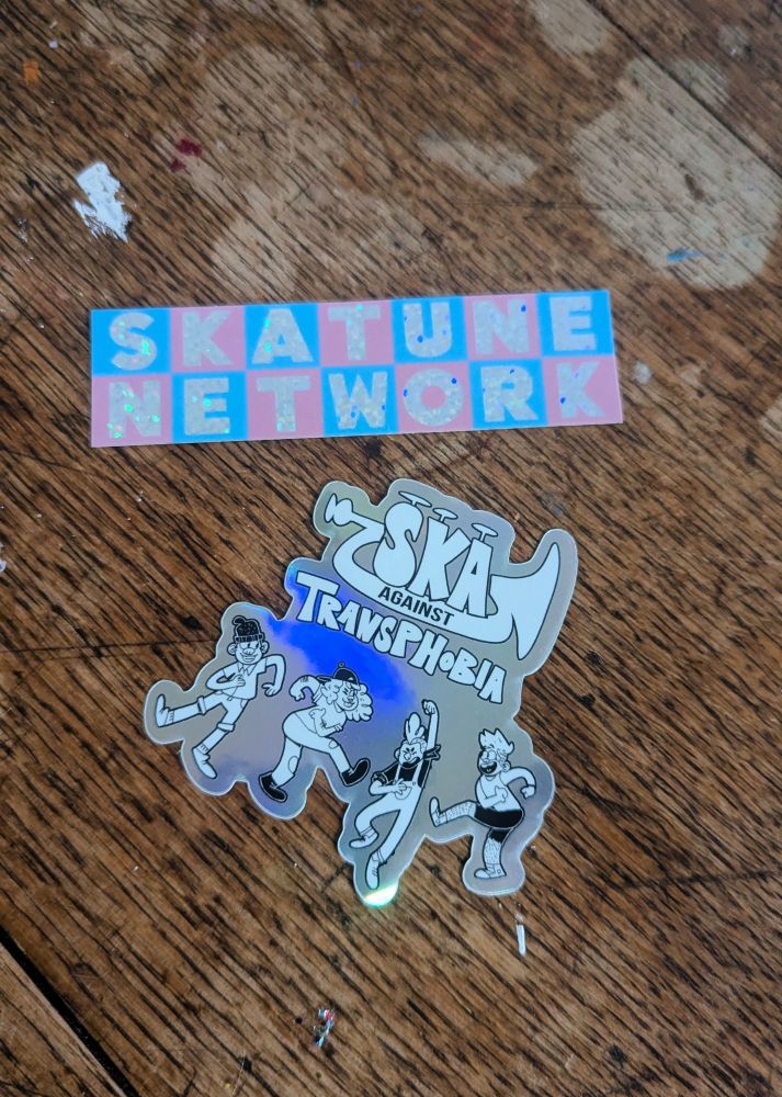 Two stickers on a wood table. Top one says Skatune Network in pride colors. The other says Ska Against Transphobia, with people dancing below it.