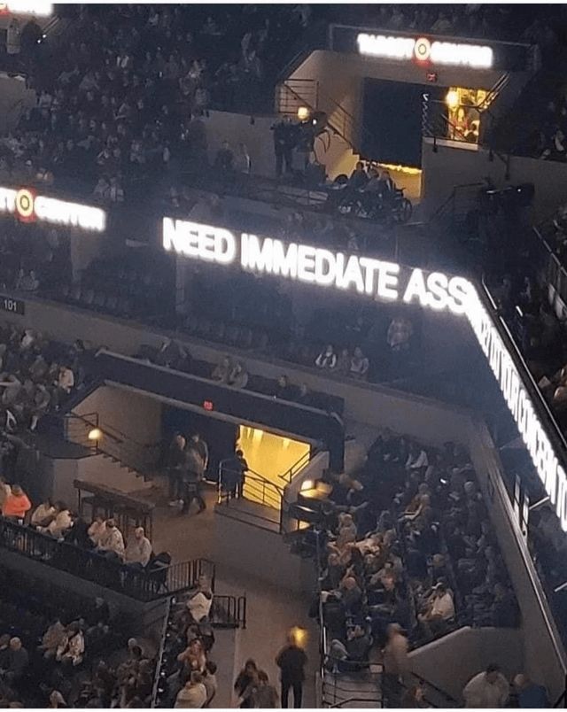 A picture of the inside seating of a stadium. There is a digital banner stretching around a corner such that you can only see the words 'Need Immediate Ass' when the message is almost surely 'need immediate assistance'