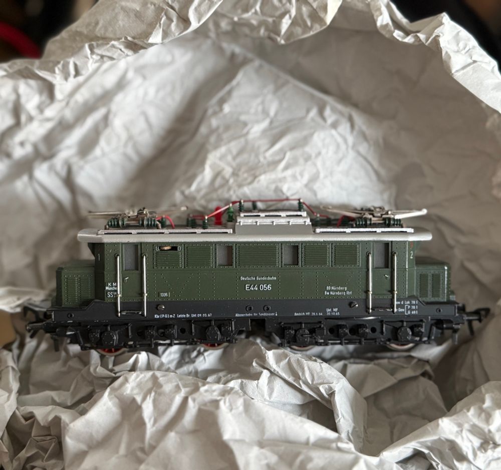 Hier ist eine tastbare, bildhafte Beschreibung des Objekts für eine blinde Person:

⸻

Du hältst ein Modell einer alten Elektrolokomotive in der Hand. Das Modell liegt in zerknülltem, weichem Packpapier, als sei es gerade ausgepackt worden.

Die Lok selbst ist:

Form & Aufbau
	•	Sie ist lang und rechteckig, ungefähr wie ein kleiner Ziegelstein, aber mit vielen feinen Anbauteilen.
	•	Auf beiden Seiten befinden sich schwarze, filigrane Drehgestelle (die Radsätze), die wie kleine technische Skulpturen wirken – viele winzige Räder, Stangen und Rohre.
	•	An den Stirnseiten ist jeweils ein Pufferbalken mit zwei runden Puffern, wie kleine Metallknöpfe.

Farbe & Oberfläche
	•	Der mittlere Wagenkasten ist tiefgrün, matt, mit feinen geriffelten Strukturen – fast wie die Oberfläche eines alten Metallwerkzeugs.
	•	Der Rahmen und die Unterseite sind schwarz, wodurch alles sehr robust und schwer wirkt.
	•	Der gesamte Eindruck ist ein wenig militärisch oder industriell, mit klaren Kanten.

Details & Beschriftung
	•	Seitlich sind viele weiße, winzige Aufdrucke zu sehen – technische Daten, Nummern, Ortsangaben.
	•	Besonders groß steht die Loknummer: „E 41 056“.
	•	Kleine silberne Stangen und Haltegriffe verlaufen vertikal und horizontal über die Seiten.

Eindruck insgesamt

Die Lok wirkt wie ein sorgfältig gefertigtes Sammlerstück – schwer, voll detaillierter Technik und mit vielen kleinen filigranen Teilen, die man fast besser ertasten als sehen könnte. Das zerknüllte Papier um sie herum lässt den Moment wirken wie das Öffnen eines wertvollen Stücks aus einer Sammlung.

.