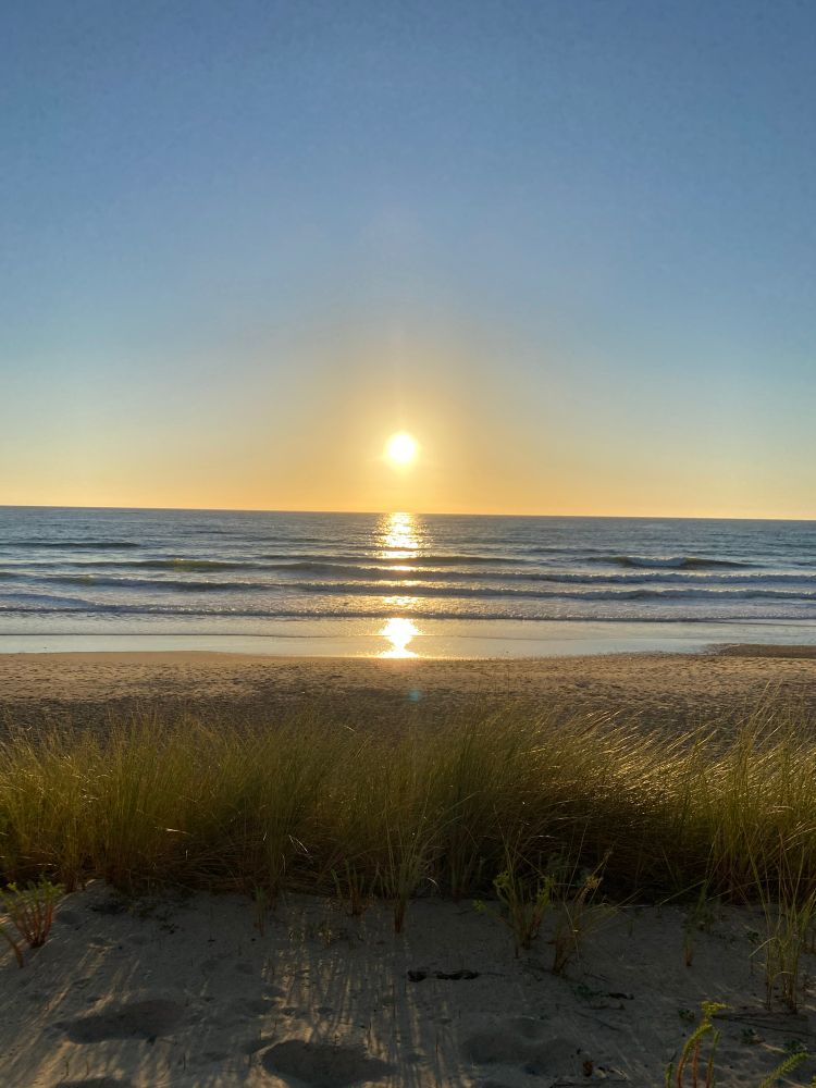 Die untergehende Sonne am Horizont spiegelt sich im recht glatten Meer. Im Vordergrund sieht man Strand mit Sand und Dünengras. Das Bild fällt durch Symmetrie und Ruhe auf.