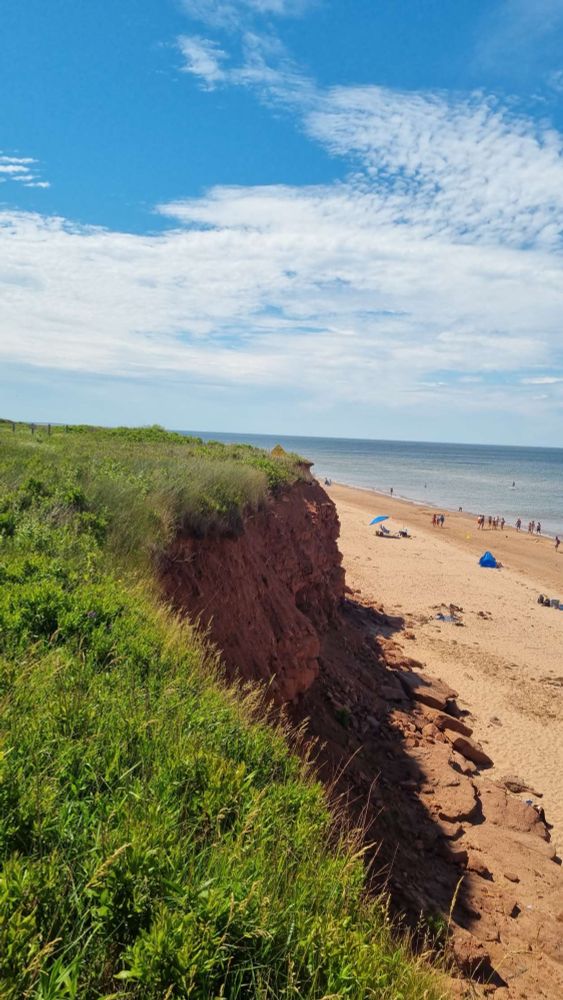 Grasfläche links fällt steil ab mit rotem Felsen zum rötlichen Strand rechts, man sieht auch Abschnitte des Atlantiks  