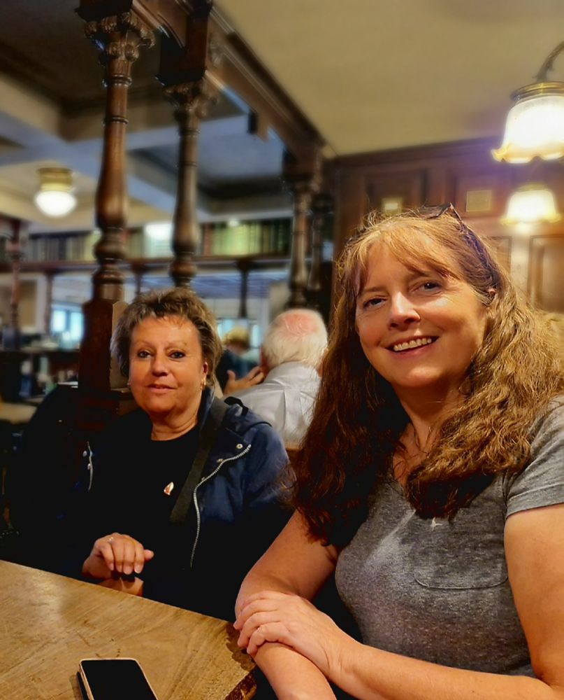 Gill and Kirsten sitting at a pub table. The pub is old-fashioned, with ornate wood pillars and panelling..