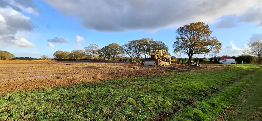 A bulldozer in the corner of a field. Earth has begun to be piled up i to a bund. Thr sky overhead is blue with grey clouds.