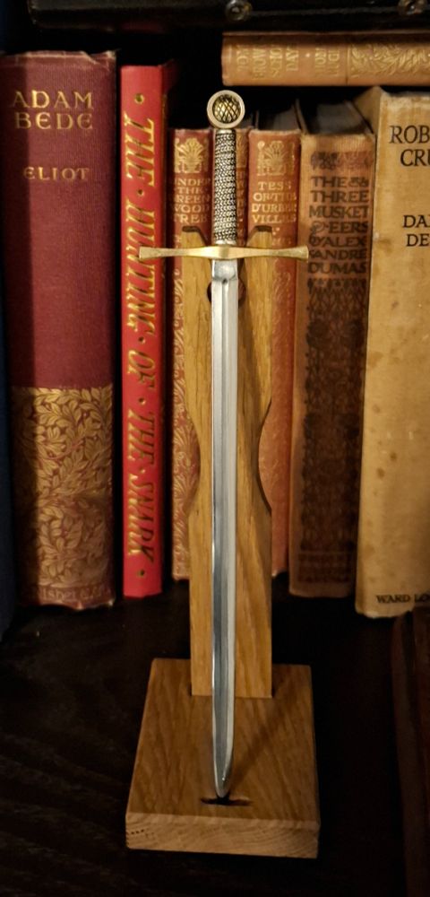 A six inch long longsword with an ornate gold coloured handle on a wooden display stand. It sits on a black bookshelf with old hardback books in the background.