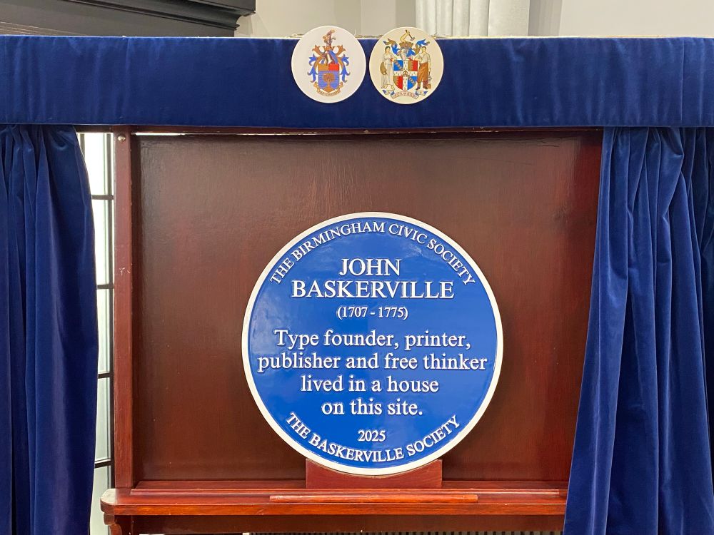A blue circular plaque on a board framed by blue curtains with two coat of arms small plaques for Birmingham and Civic Society