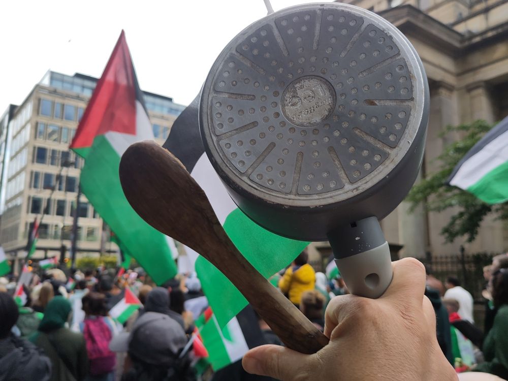 Photo of a large crowd waving Palestine flags on a city street. In the foreground a man's hand holds a saucepan and wooden spoon, in the background buildings including the neoclassical columns of Manchester Art Gallery.