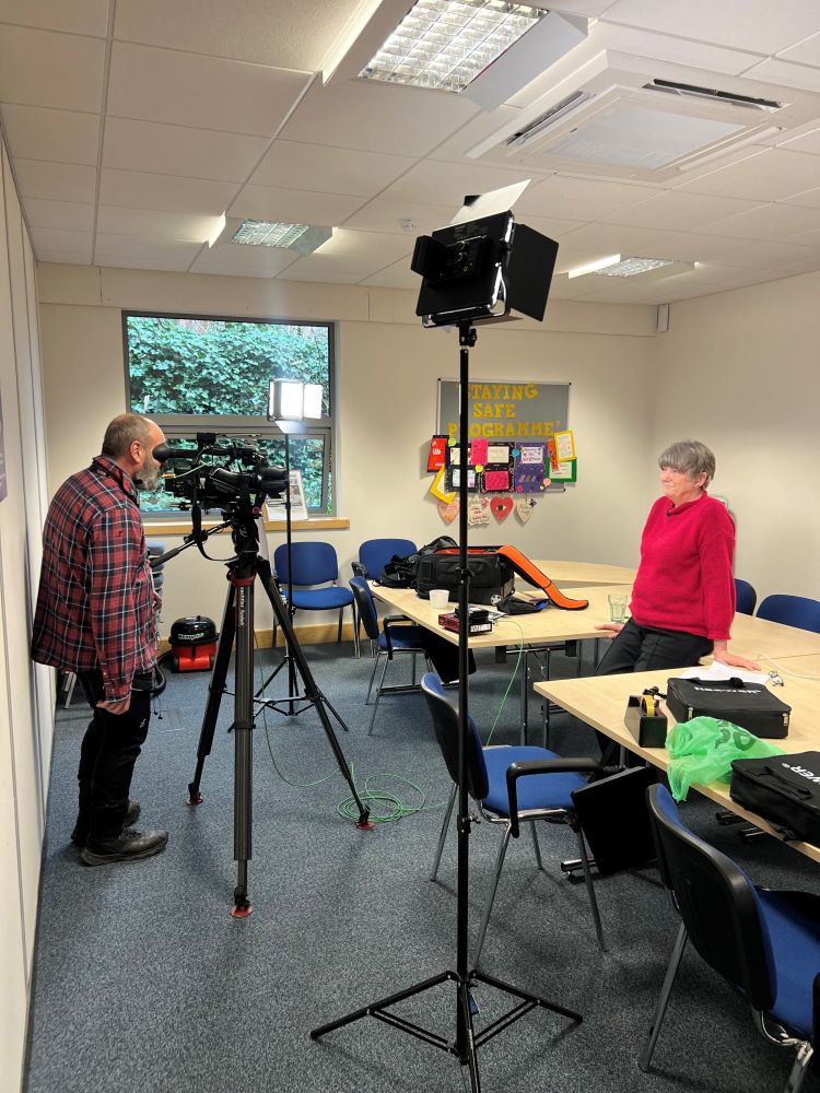 Camera man filming Janice Fife in an office, with lighting equipment