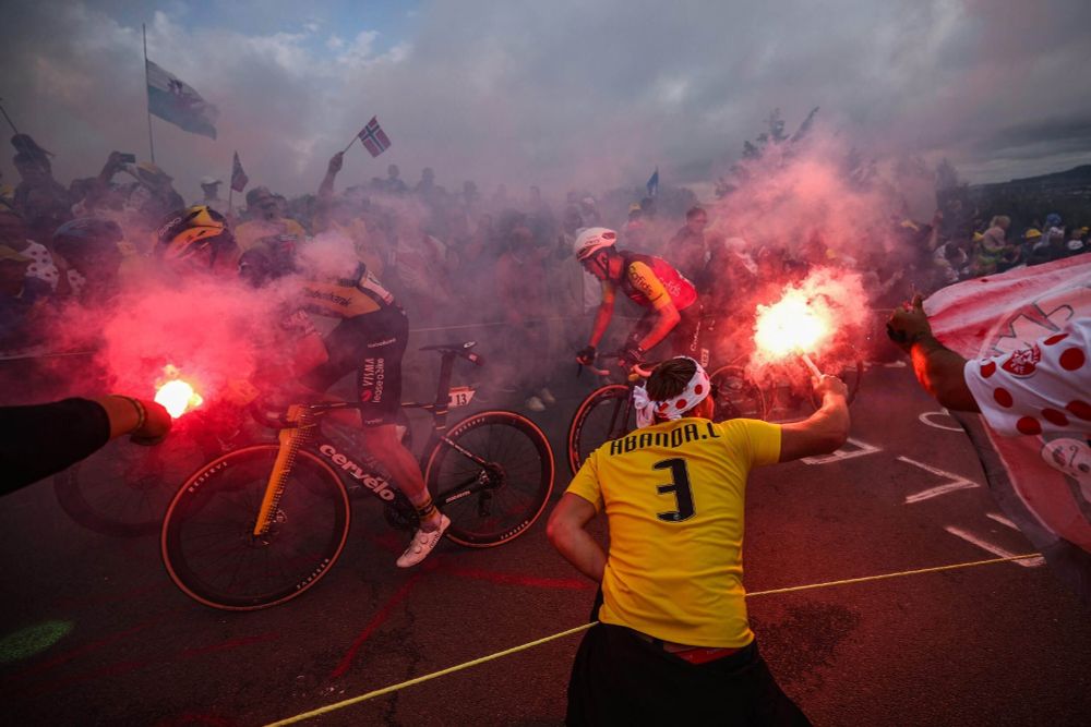 Two Tour de France cyclists in yellow and black jerseys push uphill through thick red smoke as fans crowd the roadside, waving flags and holding flares. Spectators in polka-dot gear and yellow shirts cheer passionately, creating a dramatic and intense race atmosphere.