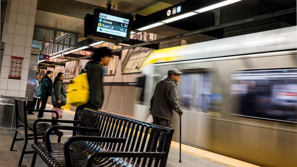 Train arriving with patrons waiting by, center is an older gent with a cane with a man with a yellow backpack that matches the train yellow pretty well.