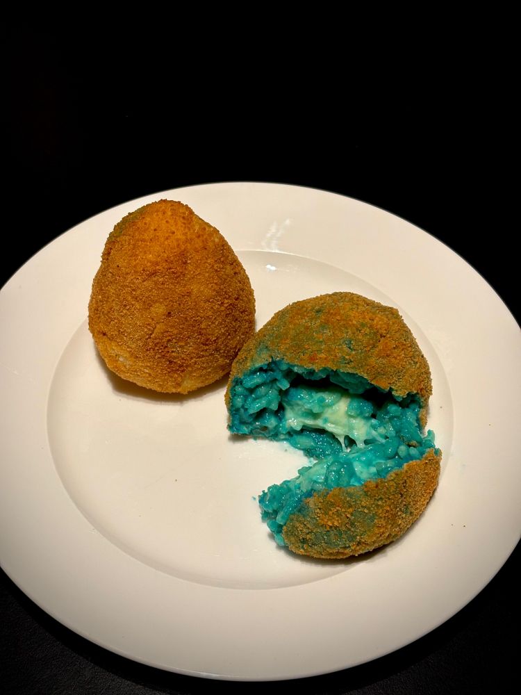 zwei arancini auf teller, einer aufgeschnitten, darin ist eine füllung aus blauem reis und mozarella