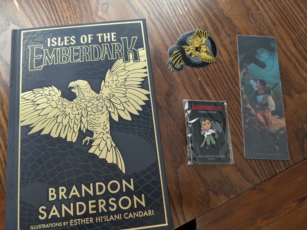The hardcover of Isles of the Emberdark. Black with gold accents, along with a matching sticker, bookmark, and character pin.