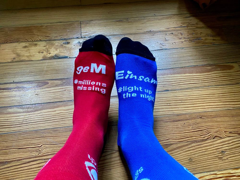 Zwei Füsse in Socken. Eine Socke ist rot und trägt die Aufschrift Millions Missing, die andere ist blau und trägt den Schriftzug light up the night.