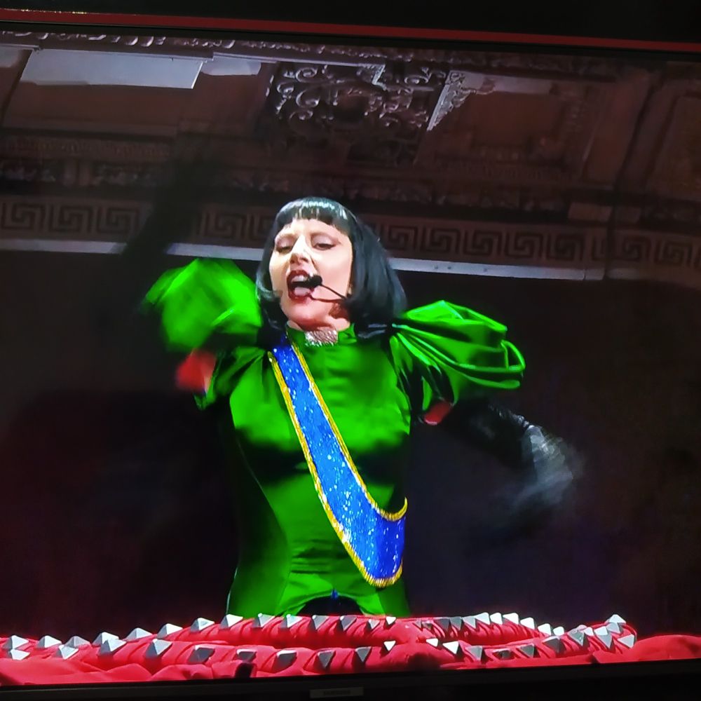 Lady Gaga aparecendo da cintura para cima usando uma peruca preta com corte channel e um vestido verde com uma faixa azul de bordas amarelas na diagonal passando pelo ombro direito.