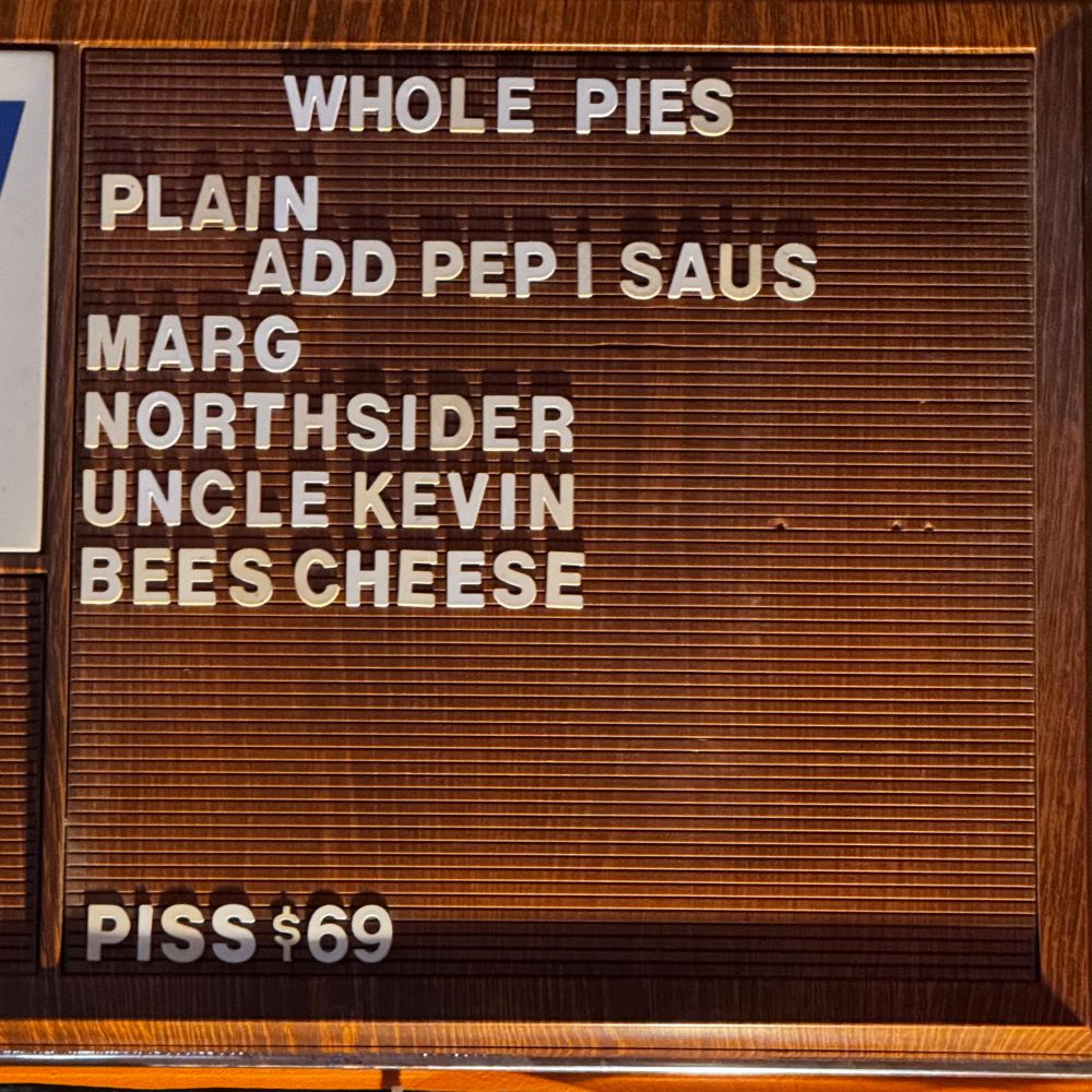 a vintage menu board that reads:

WHOLE PIES
PLAIN
ADD PEP I SAUS
MARG
NORTHSIDER
UNCLE KEVIN
BEES CHEESE
PISS $69