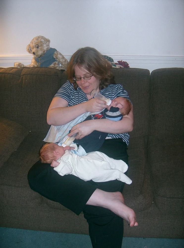 A mother of twins balancing one baby in the crook of her arm and the other baby on her lap while bottle feeding the both.