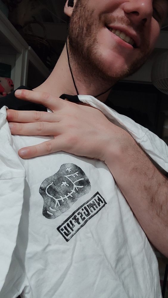 Man smiling holding a white tee shirt with black collar and black stencil logo of chiptune artist NMLSTYL