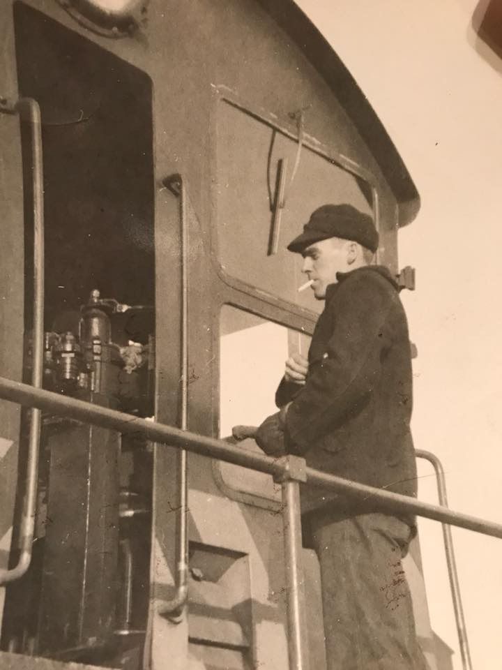 A black and white photo of a young man on the back of a locomotive engine with a cigarette in his mouth. He looks cool as hell.