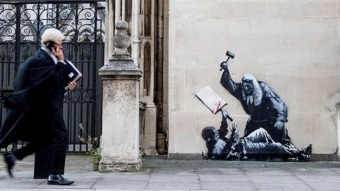 Banksy on the side of the Royal Courts of Justice in London depicts a protester being beaten down by a wigged judge holding a gavel. The protester's sign is splattered with blood. A wigged barrister walks by on the phone in the foreground.