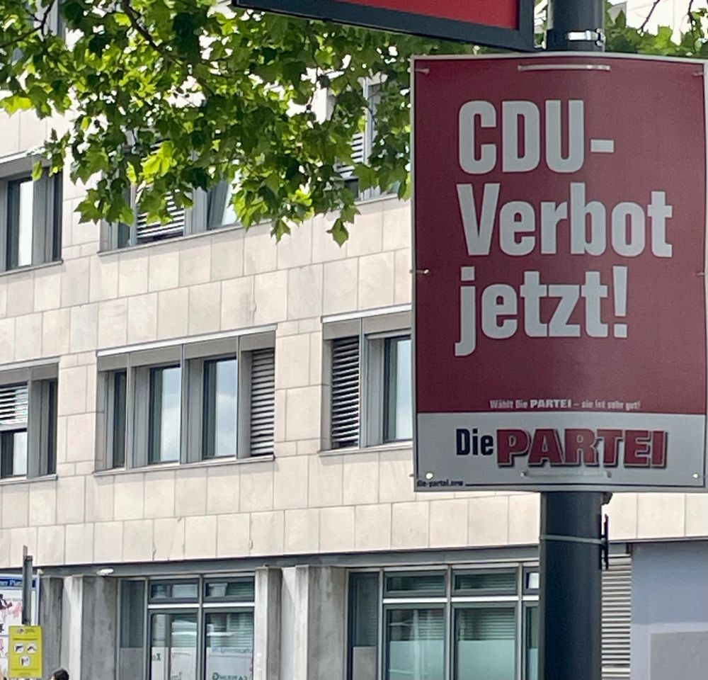 Plakat mit der Aufschrift 'CDU-Verbot jetzt!' und dem Logo der Partei 'Die PARTEI' an einem Laternenpfahl vor einem Bürogebäude
