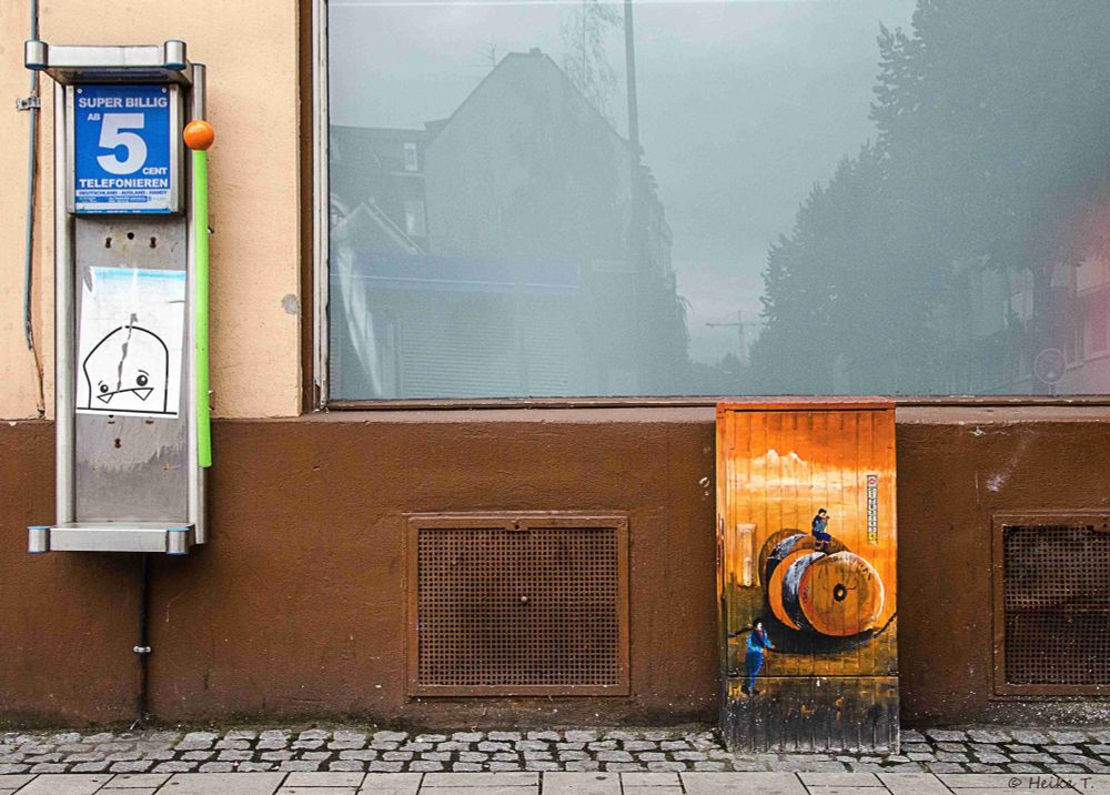 Ein Telefonautomat mit der Aufschrift 'SUPER BILLIG 5 CENT YELLOWFON' steht links. Rechts ist ein bemalter Kasten mit Kabeltrommeln und einer Person darauf.