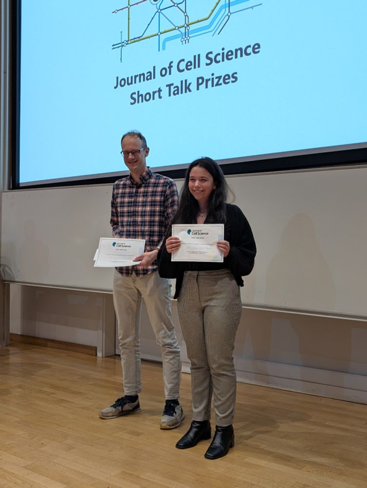 Photo of Alex Van Vliet (left) and Katy Yalci (right), holding certificates for their best talk prizes from Journal of Cell Science. The projected slide behind them says, ‘Journal of Cell Science Short Talk Prizes’, below a London Tube map.