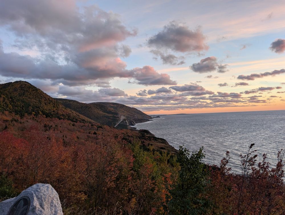 Sunset along the Cabot Trail showing sky changing colour as it touches the water. The left of the picture shows the colourful fall foliage and the winding road in the distance