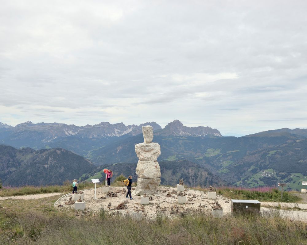 There's a mountain slope on the foreground. 3 people walk in front of stacked blocks of crystalline dolomite stone by Helmut Pizzinini. Mountain ranges are in the background.