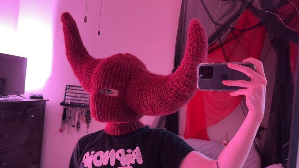 a selfie of ellen saucysphinx wearing a handmade red crochet hood, with large curved horns coming out of the sides